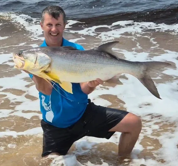Pêcheur agenouillé dans les vagues de l’océan Atlantique tenant un grand capitaine argenté capturé sur les plages d’Iguéla au Gabon.